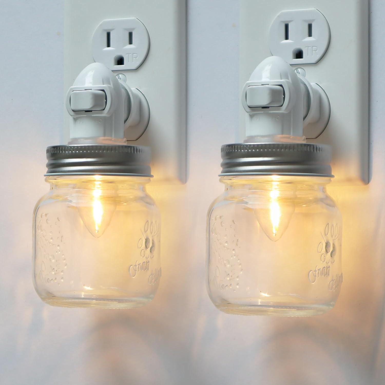 loiwon-vintage-mason-jar-plug-in-night-light-2-pack-with-on-off-switch-01 Two clear glass Mason jar night lights with brushed plastic bases, plugged into wall outlets, emitting a warm glow.
